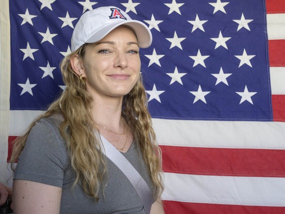 A young woman with long blonde hair and a white UArizona baseball hat smiles as she stands in front of an American flag on a wall. 