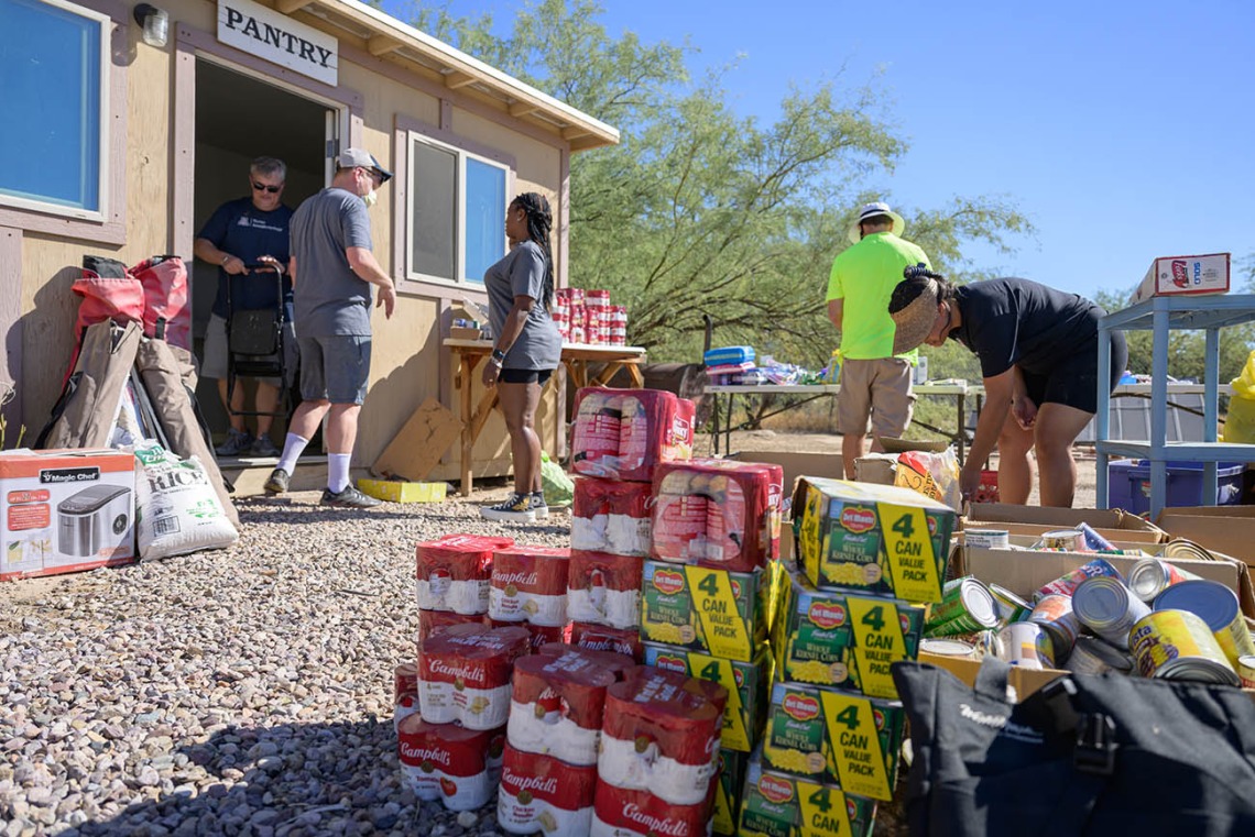 Stacks of canned goods sit on the ground outside of a small building with a sign that reads, "Pantry", while a few people outside the building are moving items into it.