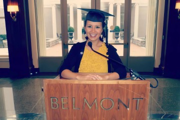 woman wearing a graduation cap and gown standing behind a lecturn that says “Belmont”